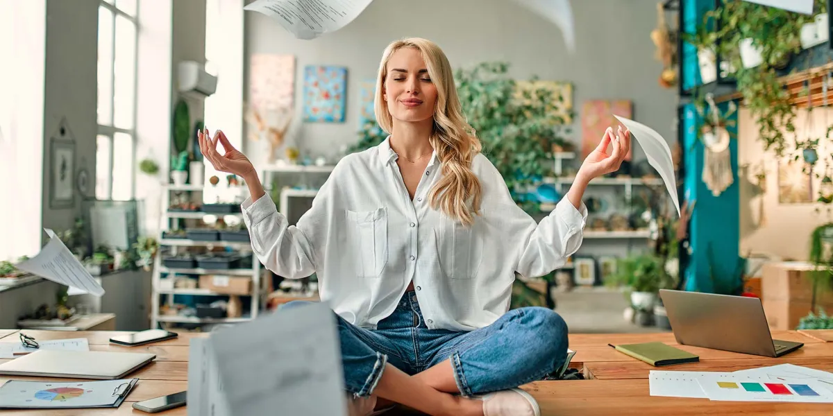 Resiliente Mitarbeiterin meditiert auf ihrem Bürotisch. Um sie herum wehen verschiedene Unterlagen. © Adobe Stock, Vasyl