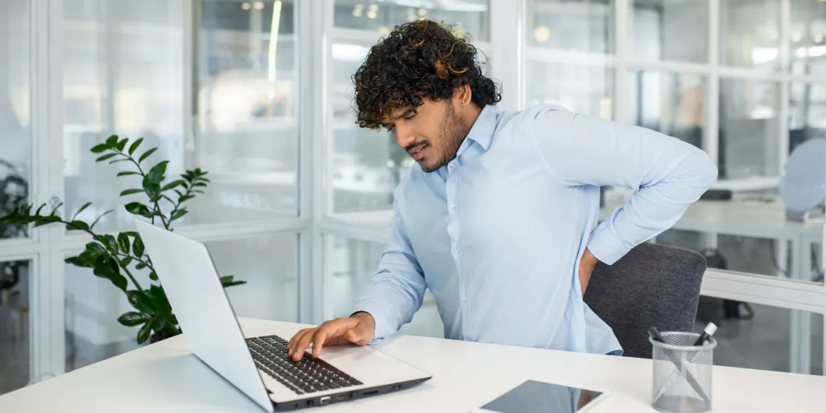 Ein junger Berufstätiger leidet unter Rückenschmerzen, während er in einem modernen Büro an seinem Laptop arbeitet. Das Bild fasst häufige Gesundheitsprobleme am Arbeitsplatz zusammen. © Adobe Stock, Liubomir